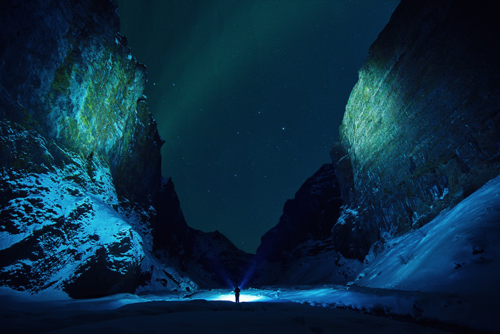 A tiny figure standing alone in a canyon at night, surrounded by towering cliffs, under the nothern lights.