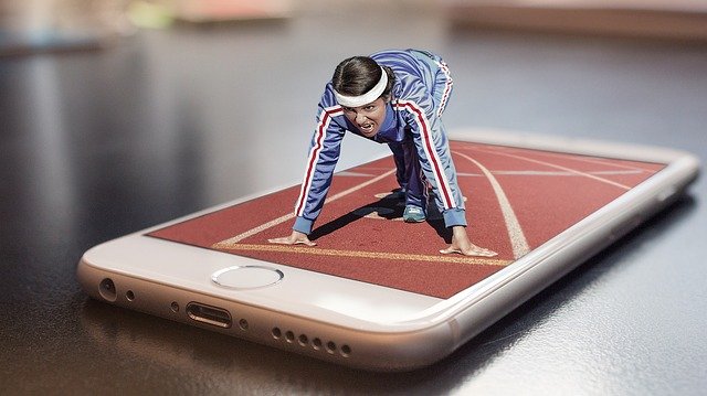 A mobile phone lies face-up on a table, with an image of a race track on the screen. A tiny, three-dimensional athlete crouched on top of the phone at the starting line of the race.