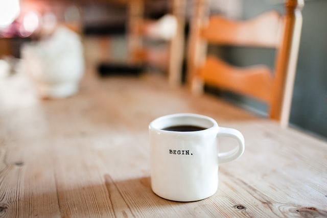 A white coffee mug sits on a wooden table; printed on it is the word "begin"